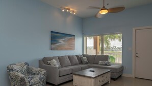 A living room with a gray sectional sofa, a matching armchair, a wooden coffee table, and a ceiling fan. A painting of a beach is on the wall, and there is a window with blinds. - Casual Condo Near Beach w/ Pools, hot tub & Grill! (South Padre Island)