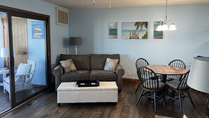 Ein Wohnzimmer mit grauem Sofa, weißem Hocker, einem Esstisch mit Stühlen und einem Deckenventilator.