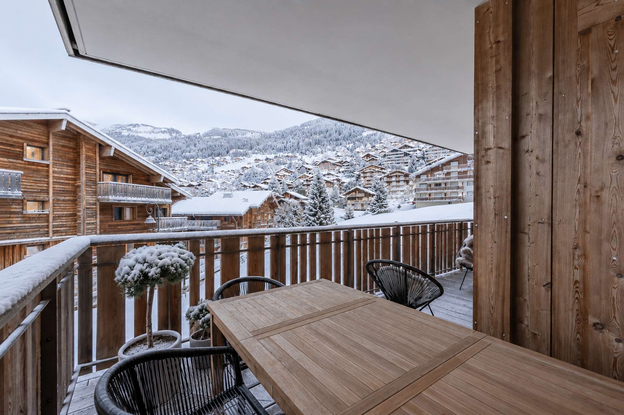 A wooden balcony with a table and chairs, overlooking a snowy mountain village.