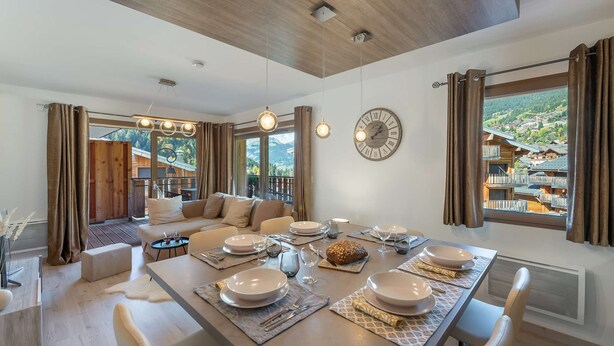 A modern dining area with a wooden table set for a meal, a sofa, and a view of a mountainous village. - Les 4 ELEMENTS - A11 (CHATEL)
