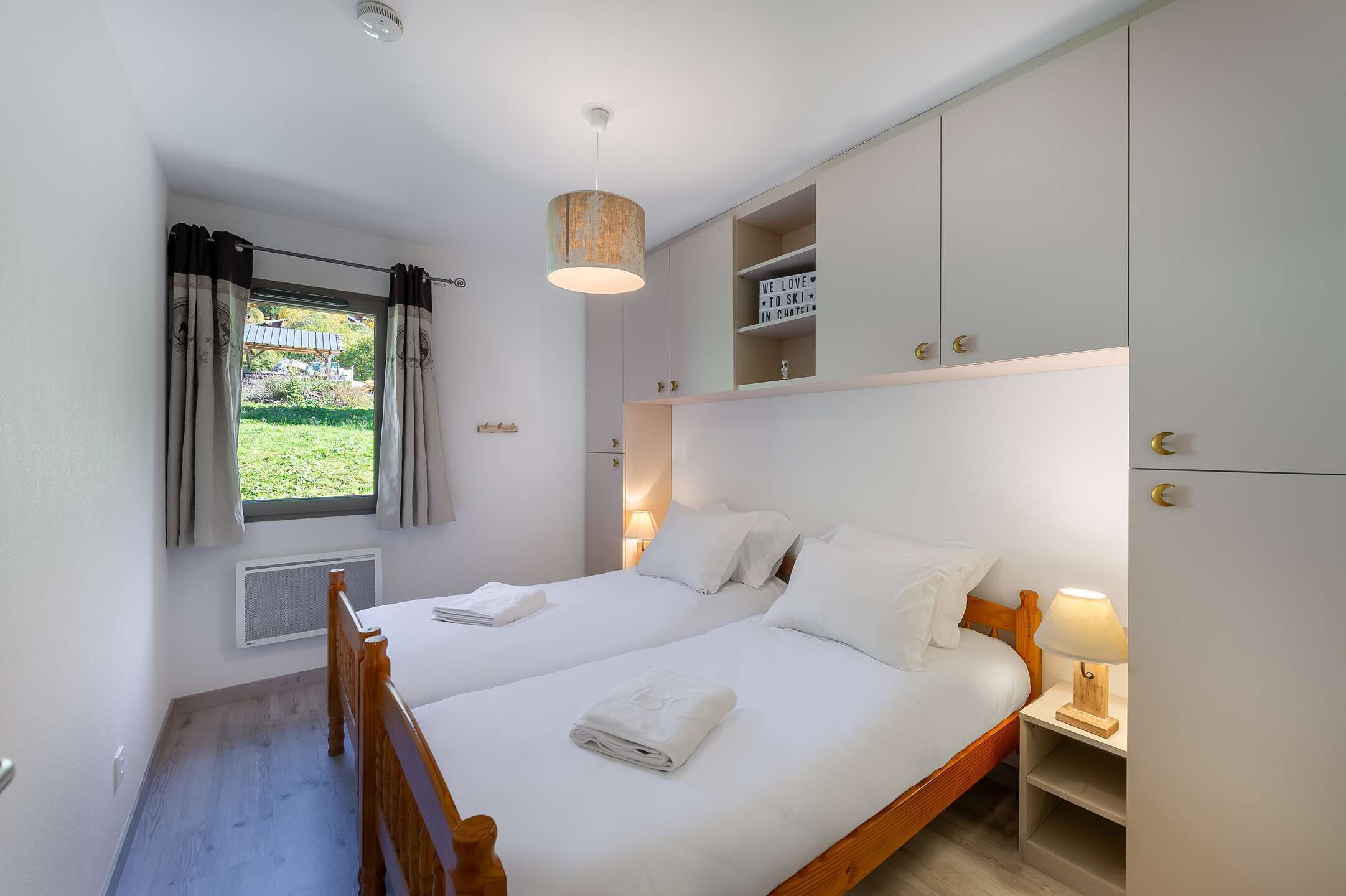 Un lit double avec du linge de lit blanc, une table de chevet avec une lampe, une armoire et une fenêtre avec des rideaux.