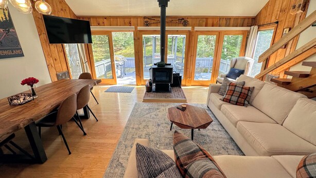 A cozy living room with a fireplace, wooden walls, a sofa, a dining table, and a TV. - 43TV peaceful retreat in the White Mountain, fireplace, fire pit, large yard, WiFi, AC! (Twin Mountain)