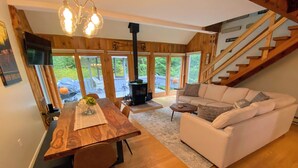 A cozy living room with a wooden dining table, white sofa, and a staircase leading to an outdoor area. - 43TV peaceful retreat in the White Mountain, fireplace, fire pit, large yard, WiFi, AC! (Twin Mountain)