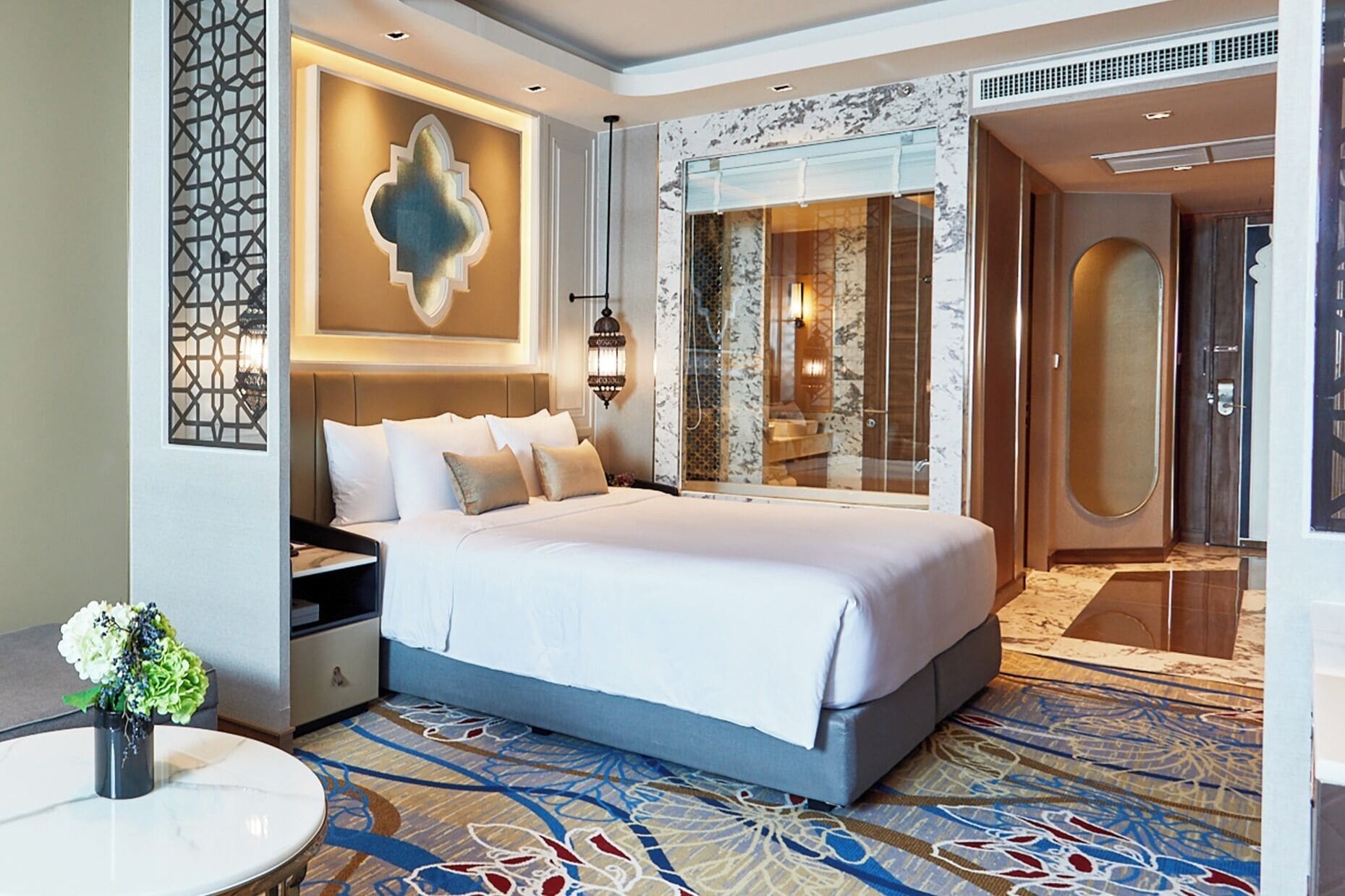 A modern hotel room with a large bed, a nightstand, a round table with a vase of flowers, and a bathroom with a glass shower enclosure.