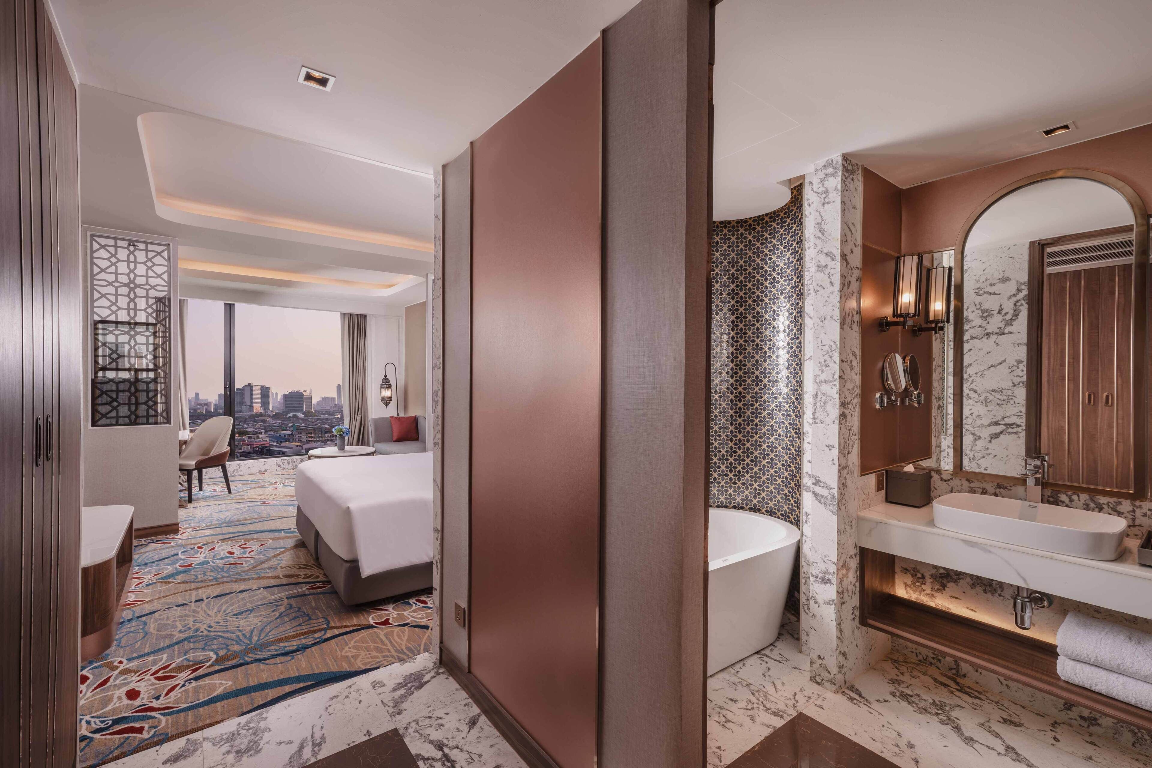 A modern hotel room with a large bed, a sitting area with a view, a bathroom with a bathtub, and a marble-tiled wall.