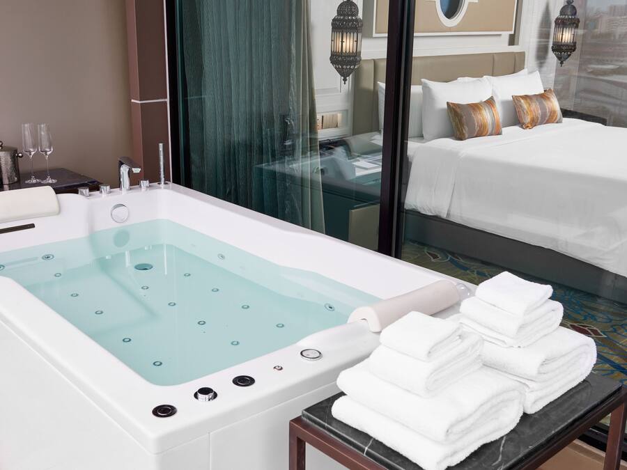A modern hotel room with a large bathtub, a neatly made bed, and a glass-enclosed shower area.