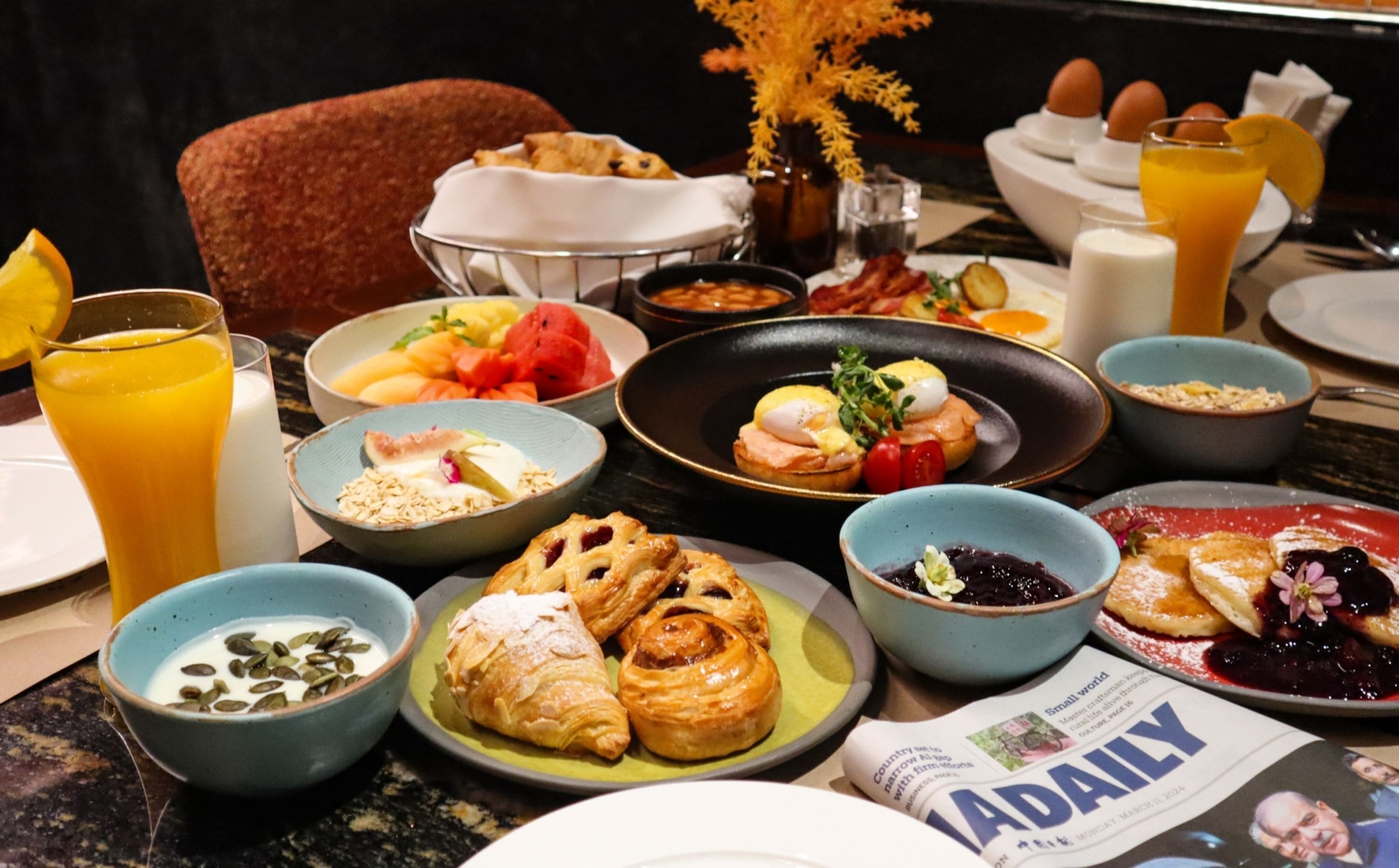 A breakfast buffet with various dishes, including pastries, fruits, and a newspaper.