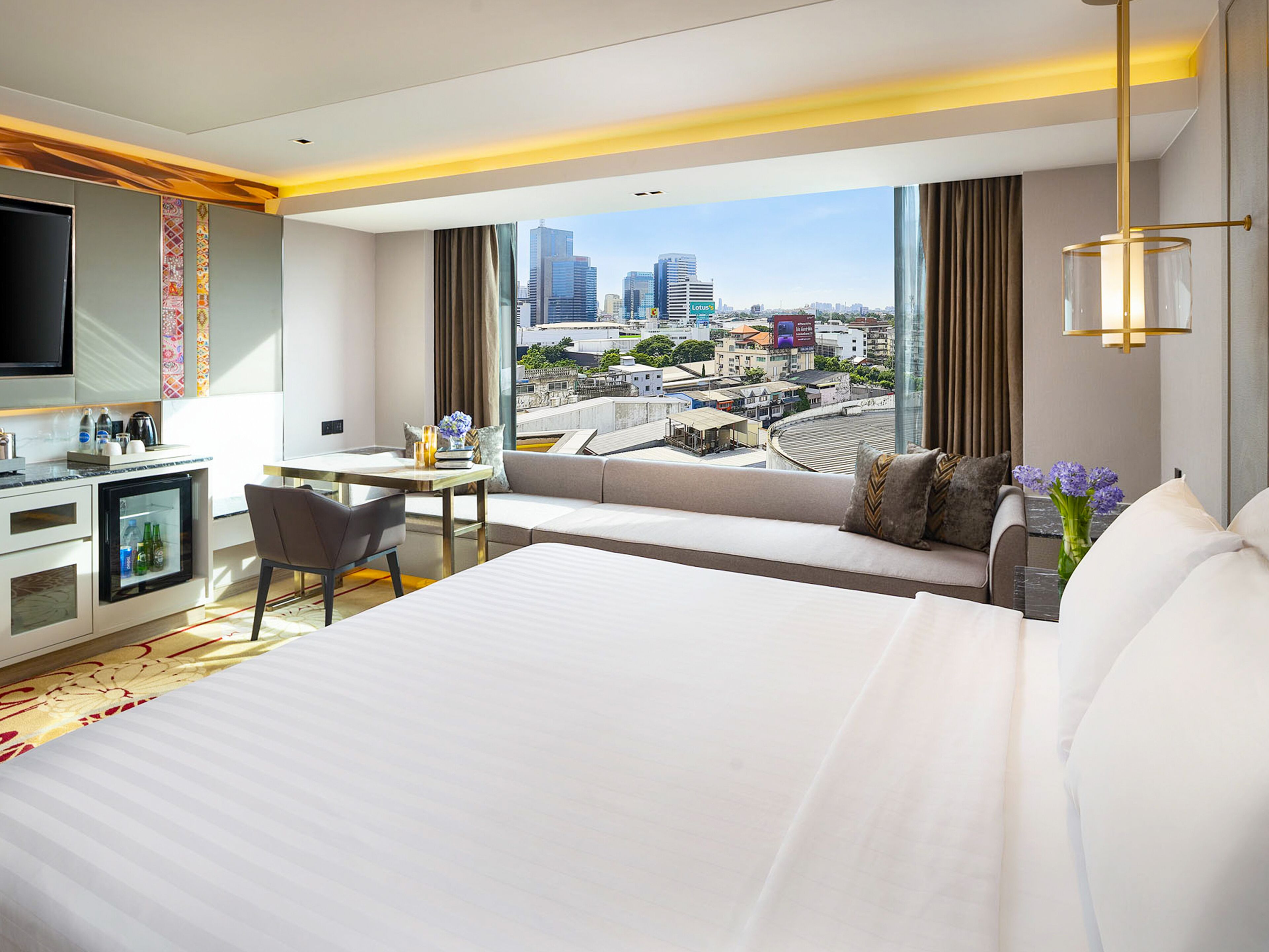 Una habitación de hotel moderna con una cama grande, un escritorio, una silla, un minibar y un televisor. La habitación tiene vistas a la ciudad a través de amplios ventanales.