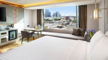 A modern hotel room with a large bed, a desk, a chair, a minibar, and a TV. The room has a city view through large windows.