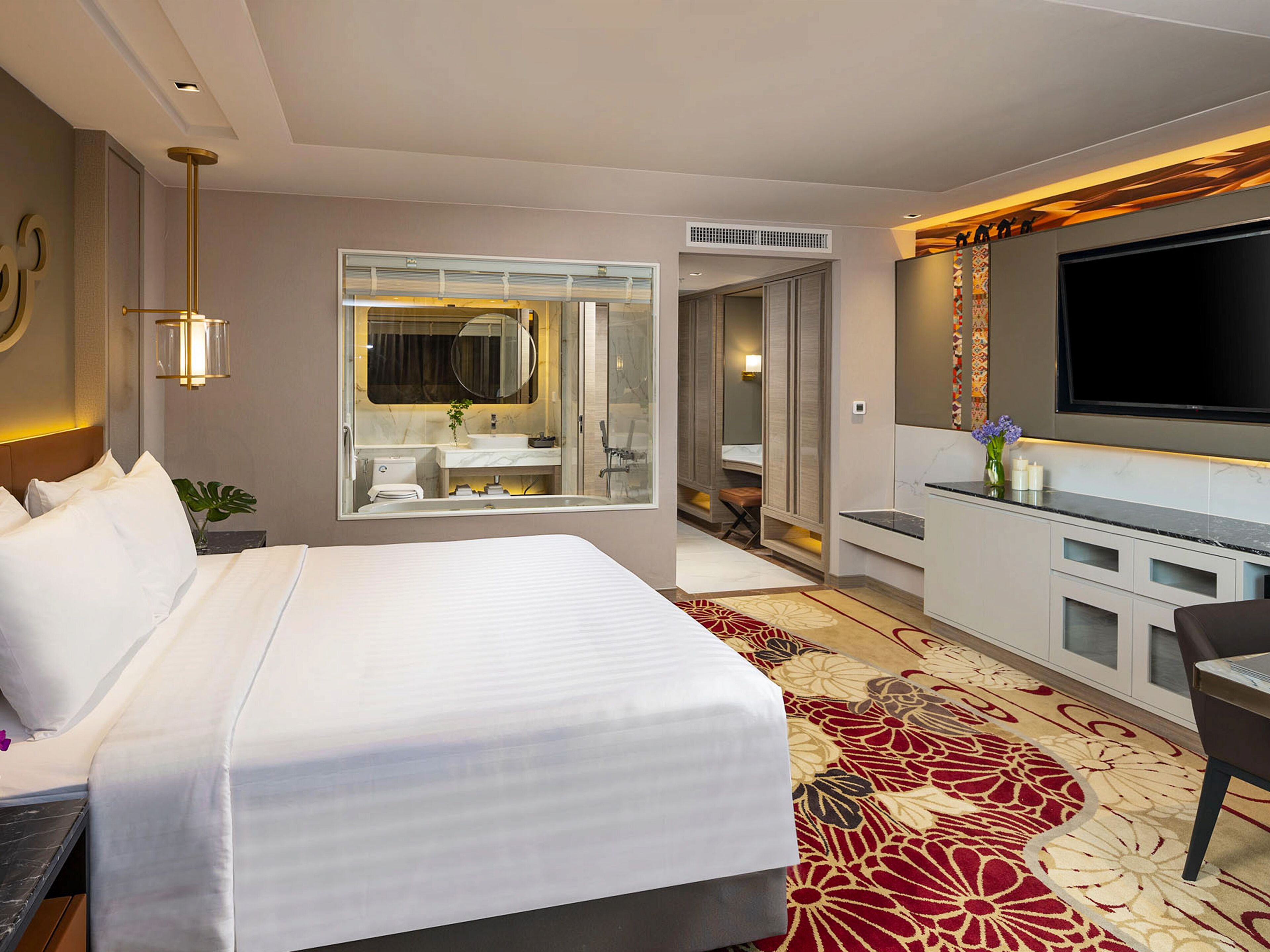 A hotel room with a large bed, a TV, a bathroom with a mirror, and a carpeted floor.