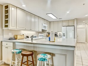 A modern kitchen with white cabinetry, a central island with stools, a microwave, and a refrigerator. - Wonderful Ocean Views, Heated Pool Access (Tybee Island)