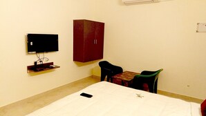 A small, simple room with a bed, a TV, a red cabinet, and a small table with a chair. - PPH Living Royal Pride (Madurai)