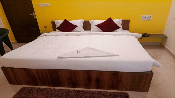 A neatly made bed with white linens and two red pillows, a wooden headboard, and a small bedside table with a phone and a bottle. - PPH Living Royal Pride (Madurai)
