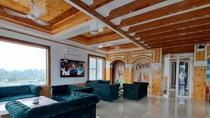 A spacious room with wooden beams, a large TV, and a 'Cheers!' mural. - ABHILASHI INN (Balh)
