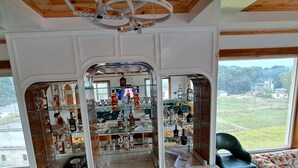 A room with a glass display of liquor bottles and glasses, a black chair, and a view of a green landscape through large windows. - ABHILASHI INN (Balh)