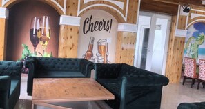 A cozy lounge area with plush green sofas, a wooden coffee table, and a mural with glasses and a 'Cheers!' sign. - ABHILASHI INN (Balh)