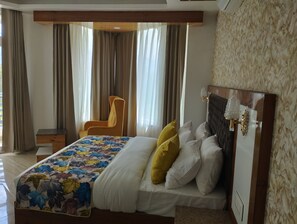 A hotel room with a bed, a chair, a nightstand, and a large window with curtains. - ABHILASHI INN (Balh)