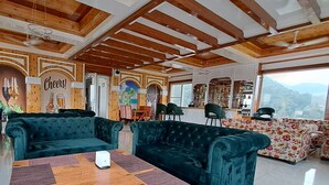 A room with a wooden ceiling, a green sofa, a wooden table, and a view of mountains through large windows. - ABHILASHI INN (Balh)