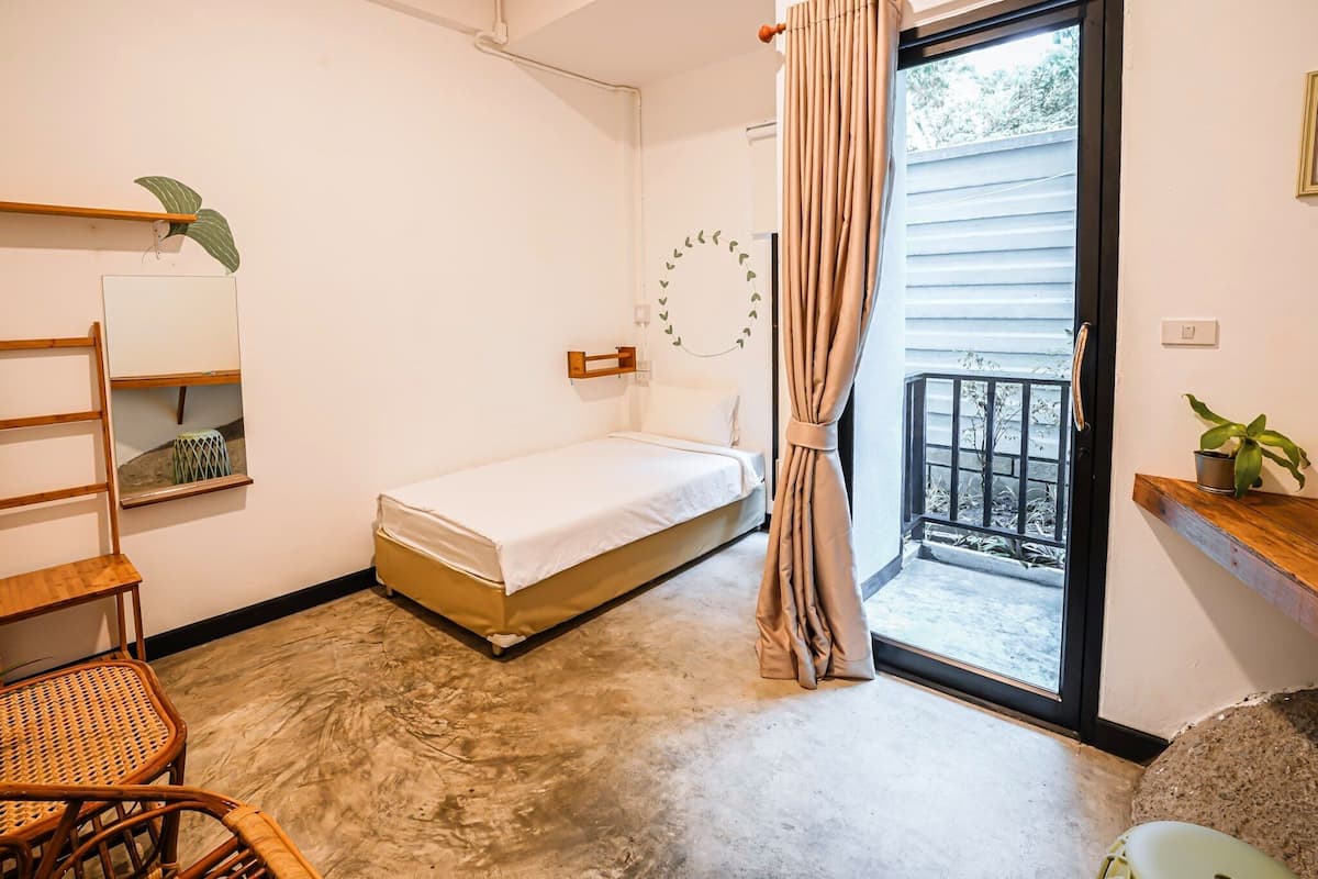 Ein Schlafzimmer mit einem Bett, einem Holzstuhl, einem kleinen Tisch, einem Spiegel und einem Balkon mit Vorhang.