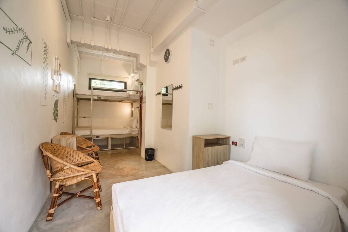 Ein schlicht eingerichtetes Hotelzimmer mit Etagenbett, einem Einzelbett, einem Korbstuhl und einem kleinen Holzschrank.