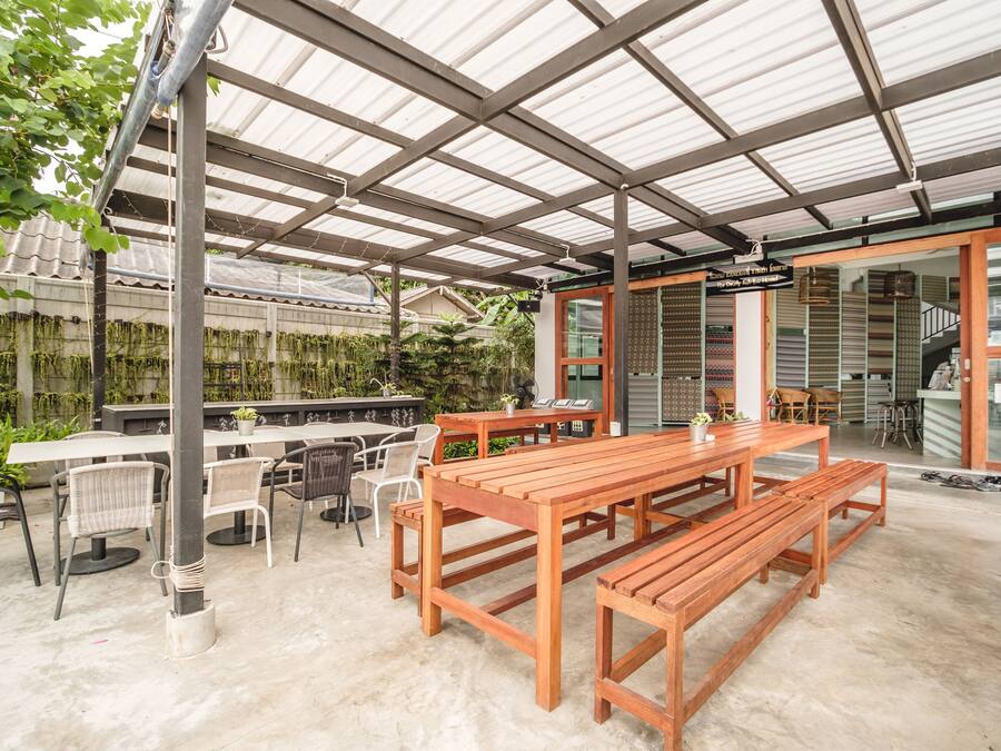 An outdoor patio area with a wooden table and benches, surrounded by chairs, under a metal roof structure.