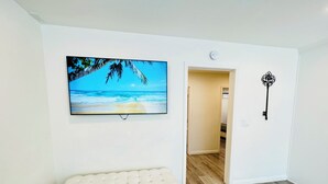Una habitación con un banco blanco, un televisor mostrando una escena de playa, una puerta que da a otra habitación y un llavero en la pared.