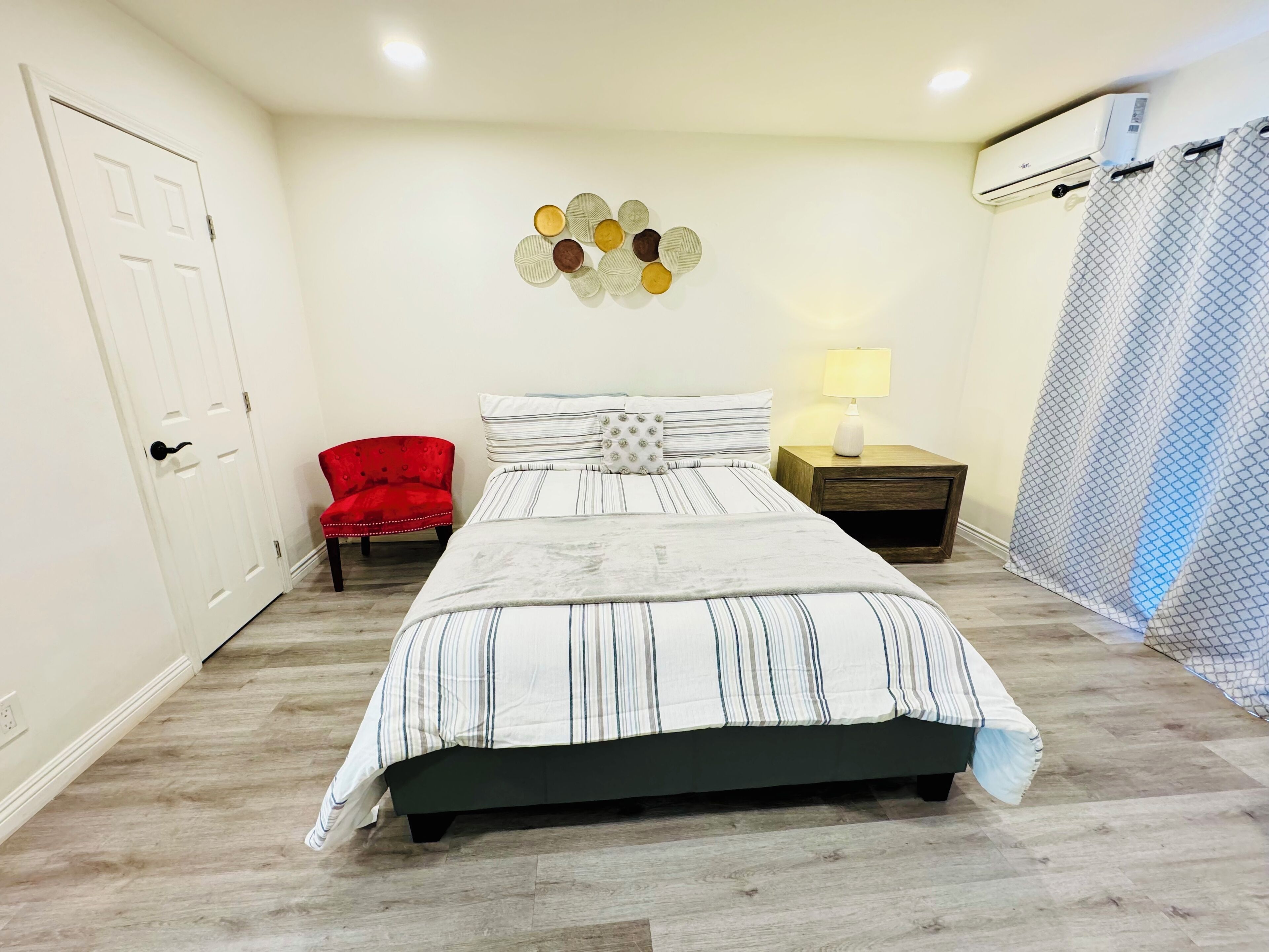 Une chambre à coucher avec un lit, une chaise rouge, une table de chevet avec une lampe, un climatiseur fixé au mur et un rideau.