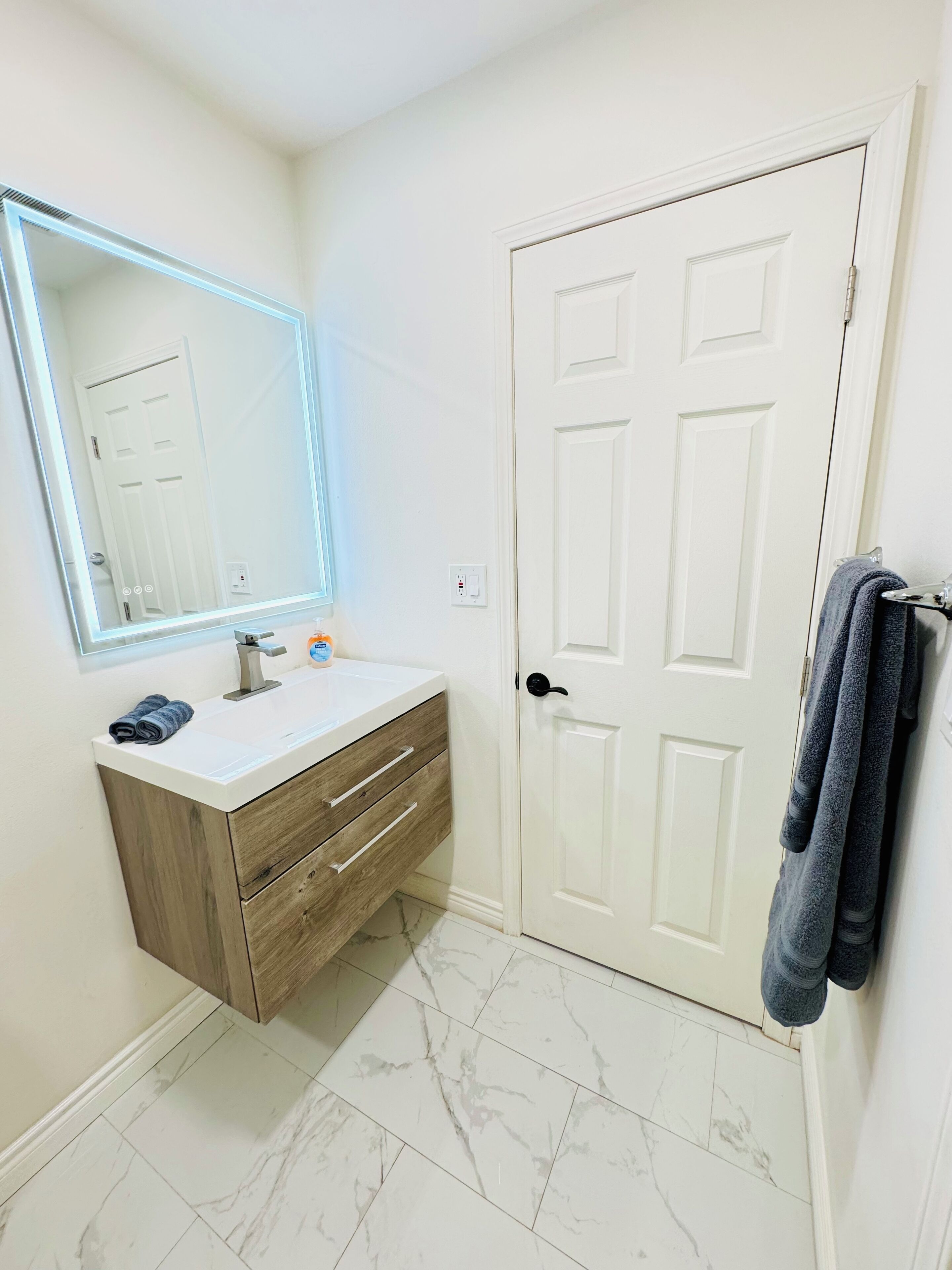 A bathroom with a white vanity, a mirror with LED lighting, a white door, and a towel rack with towels.