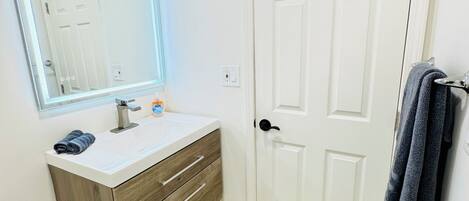 A bathroom with a white vanity, a mirror with LED lighting, a white door, and a towel rack with towels.