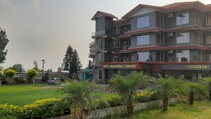 A multi-story hotel with a restaurant on the ground floor, surrounded by a well-maintained lawn and various trees. - HOTEL DHARAMSHALA PARADISE (Dharamshala)
