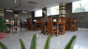 A spacious dining area with wooden chairs and tables, large windows, and a stone wall. - HOTEL DHARAMSHALA PARADISE (Dharamshala)
