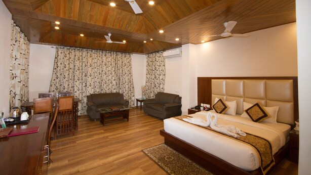A hotel room with a wooden ceiling, a large bed with a headboard, two sofas, a dining table, and a ceiling fan. - HOTEL DHARAMSHALA PARADISE (Dharamshala)
