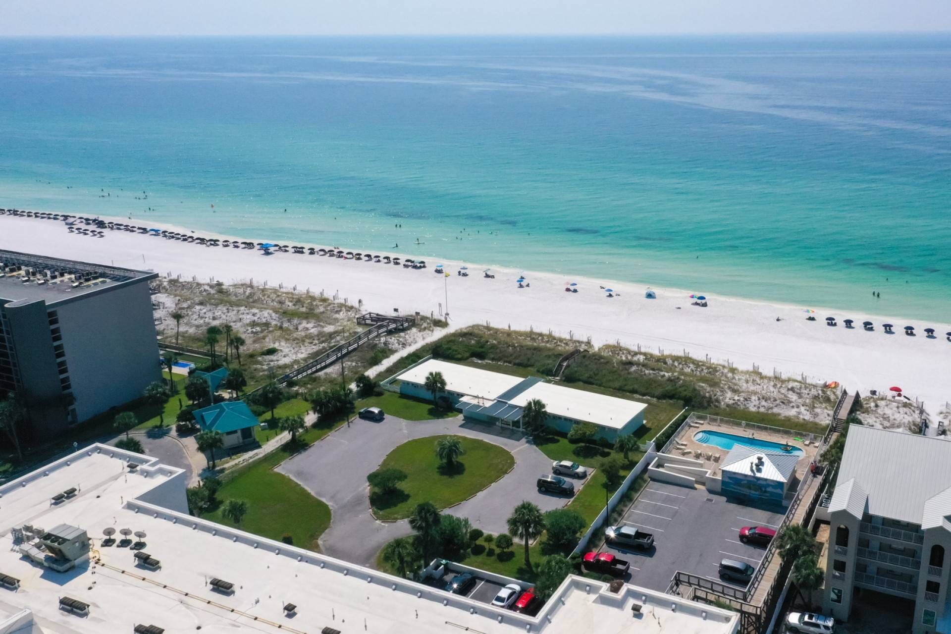 Vista aérea de un hotel frente al mar, con playa, estacionamiento y piscina.