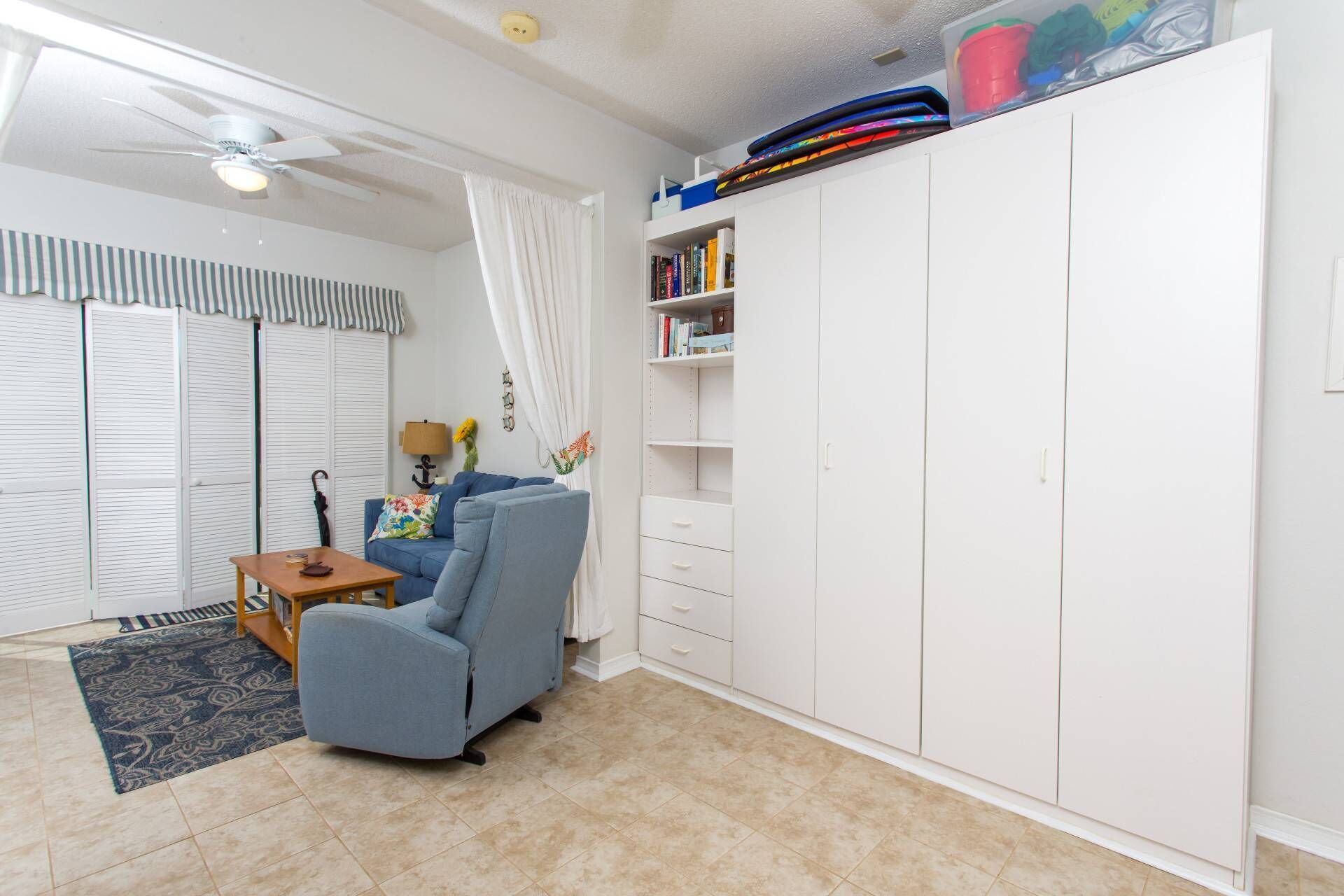 Une pièce avec un fauteuil bleu, une table en bois, une armoire blanche et un ventilateur de plafond.