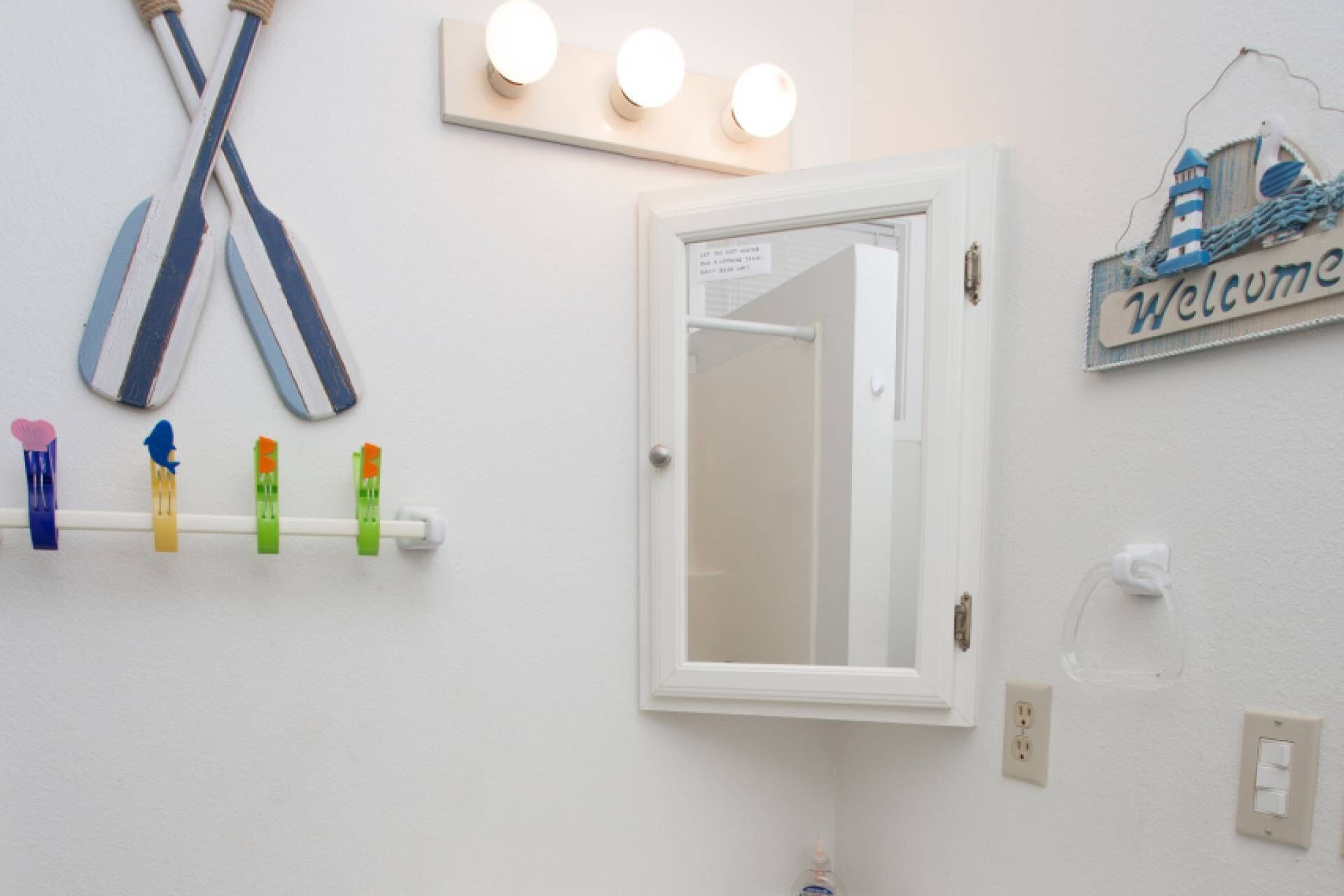 A bathroom with a white door, a towel rack with colorful clothespins, a welcome sign, and a light fixture above the door.