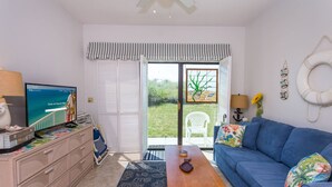 Una sala de estar con un sofá azul, una mesa de centro de madera, un televisor sobre un aparador y vista a un jardín por una puerta corrediza de vidrio.