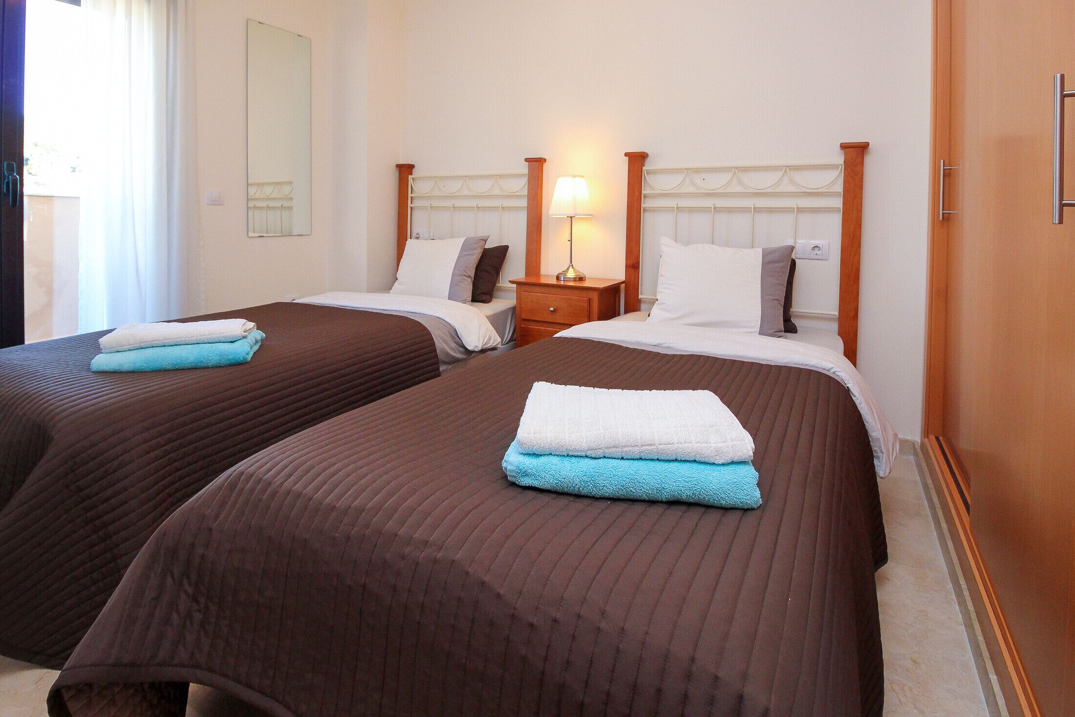 A hotel room with two beds, each with a wooden headboard, a bedside table with a lamp, and a mirror on the wall.