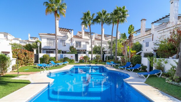 A swimming pool with lounge chairs, palm trees, and white buildings with tiled roofs.