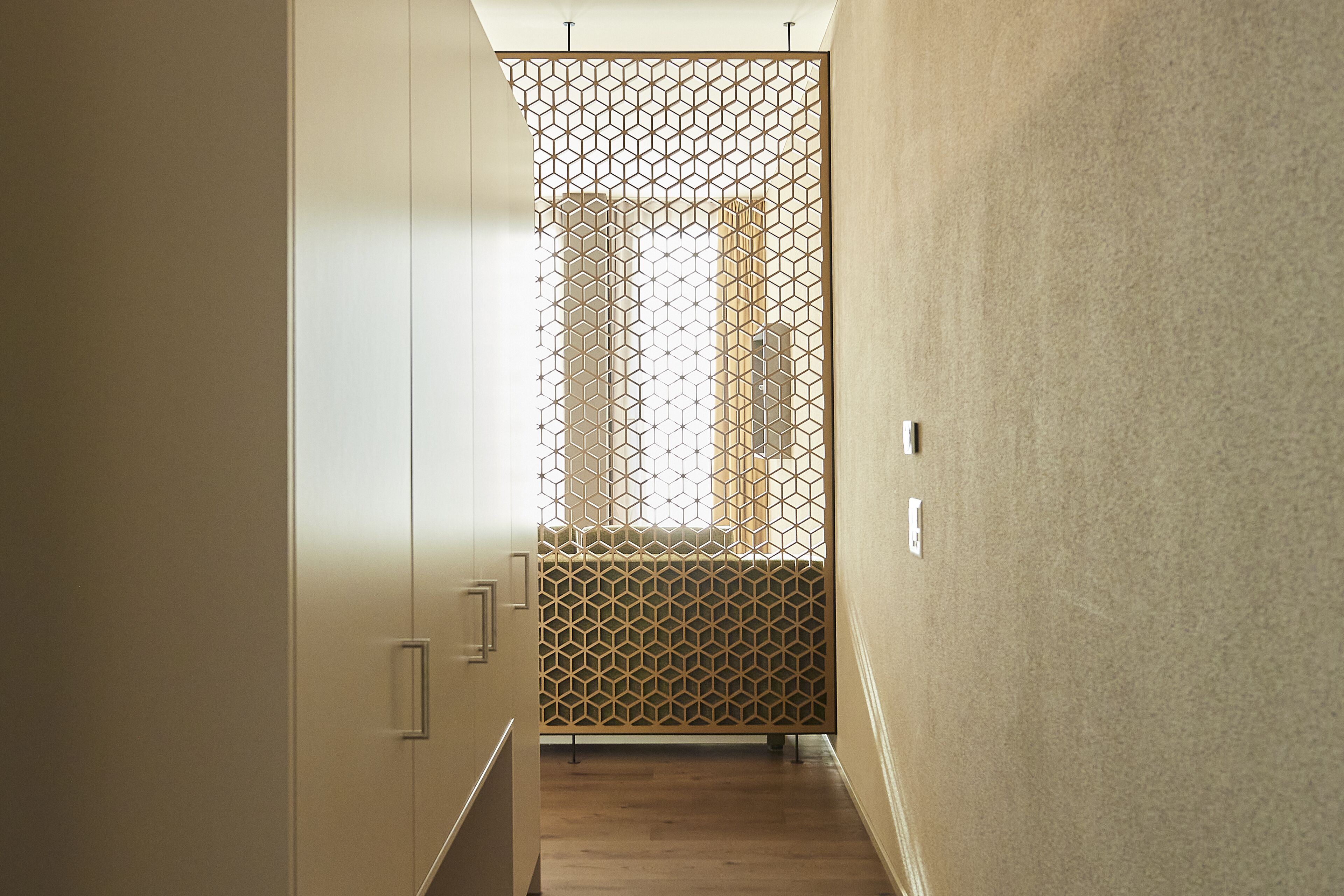 A narrow hallway with a geometric patterned partition, wooden flooring, and a view of a building through a window.