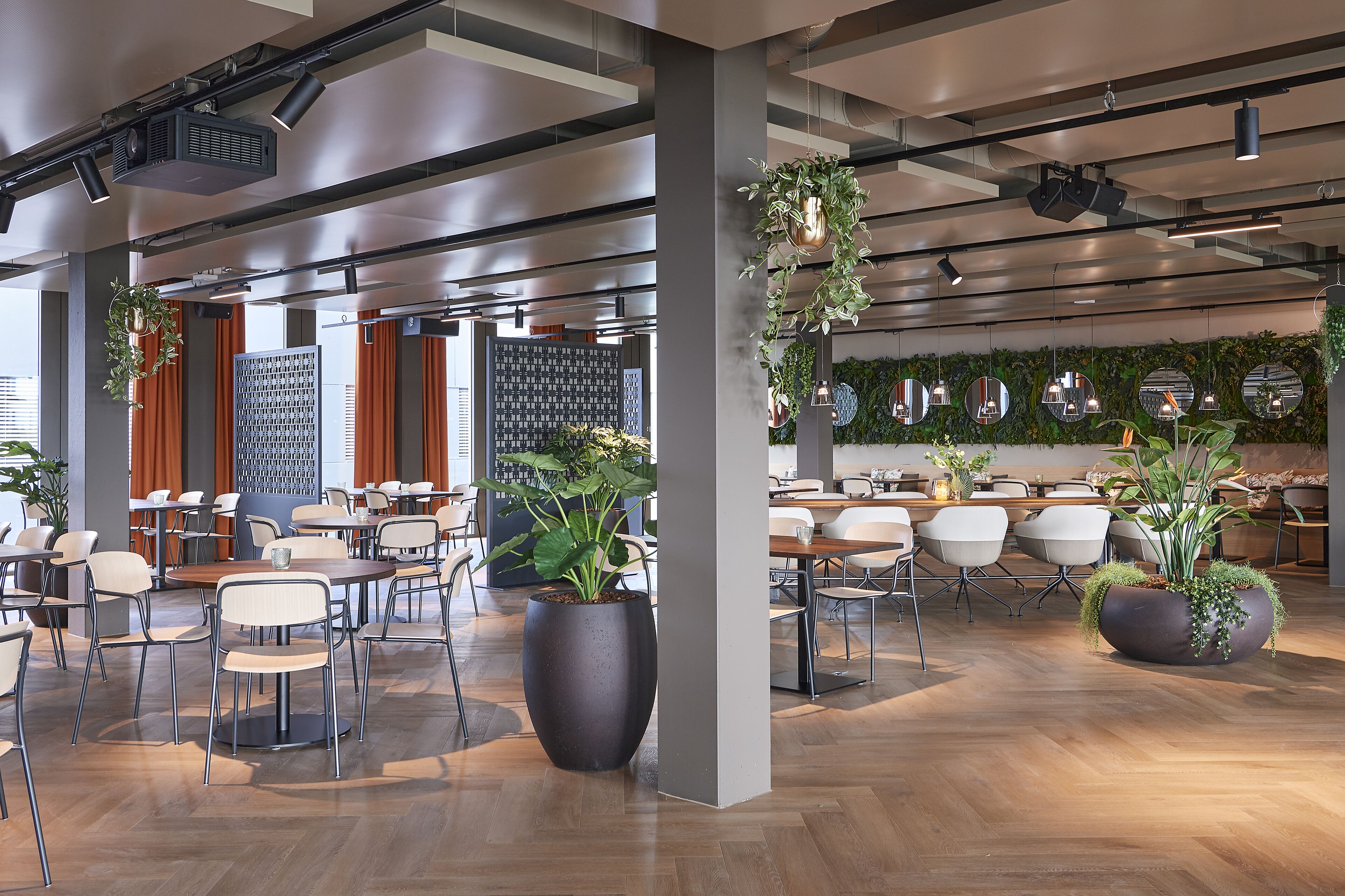 A spacious indoor area with multiple tables and chairs, large potted plants, and a modern ceiling design with exposed beams and lighting fixtures.