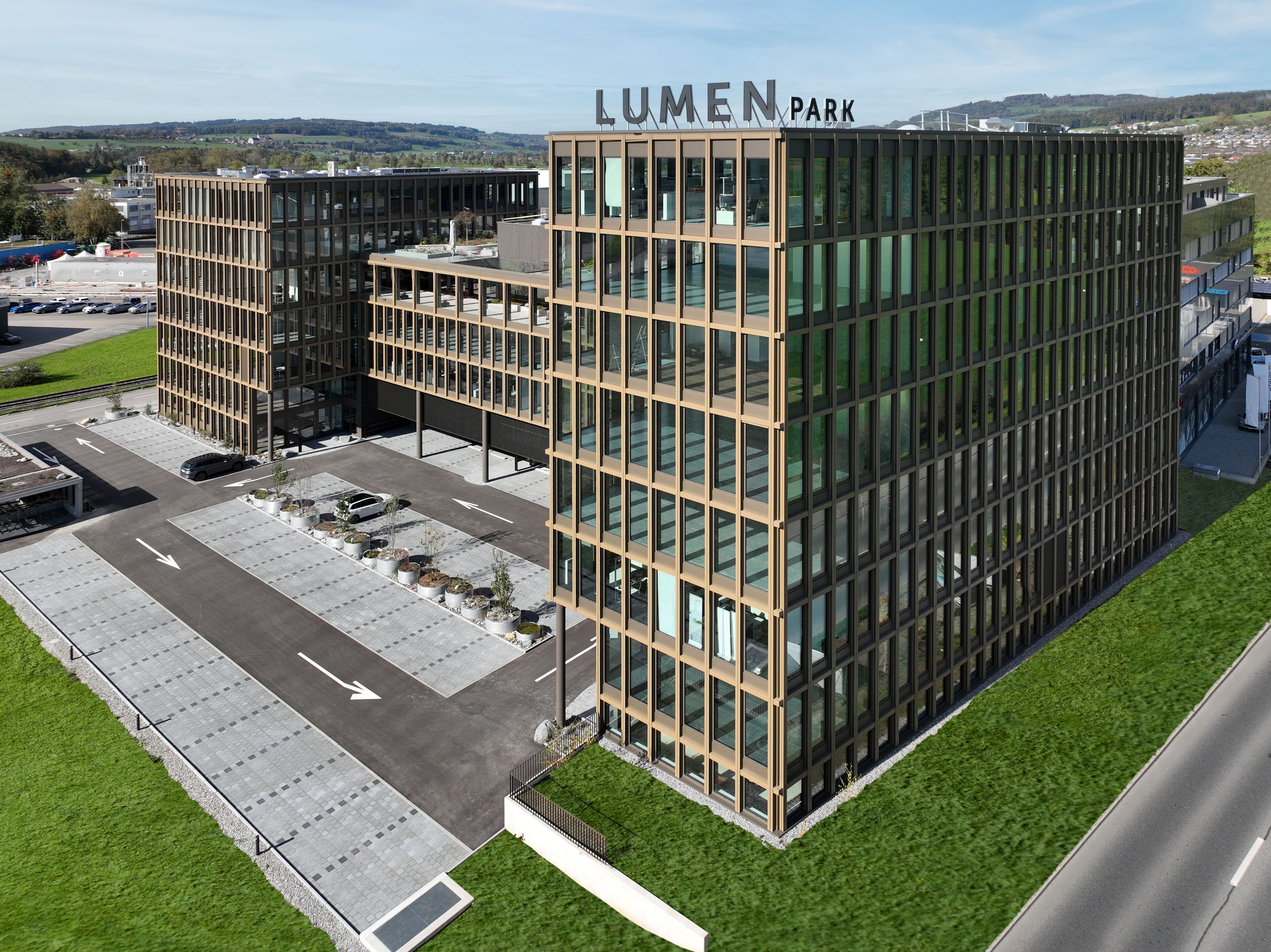 Ein modernes Gebäude mit der Aufschrift "Lumen Park" auf dem Dach. Es verfügt über einen Parkplatz mit Pfeilmarkierungen und eine grüne Rasenfläche.