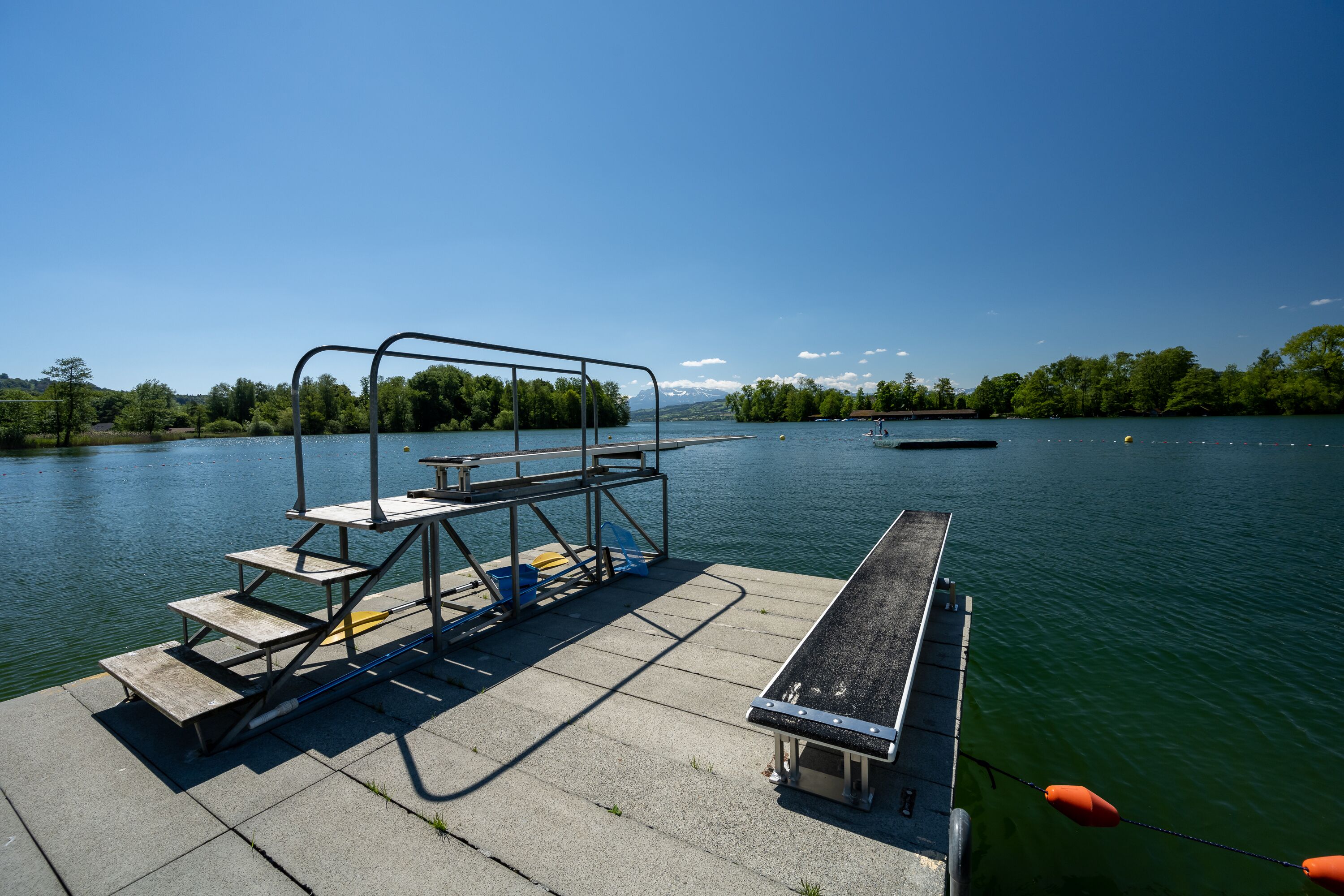 Un muelle de concreto con una escalera de metal y escalones que dan a una masa de agua, rodeado de árboles y un cielo despejado.