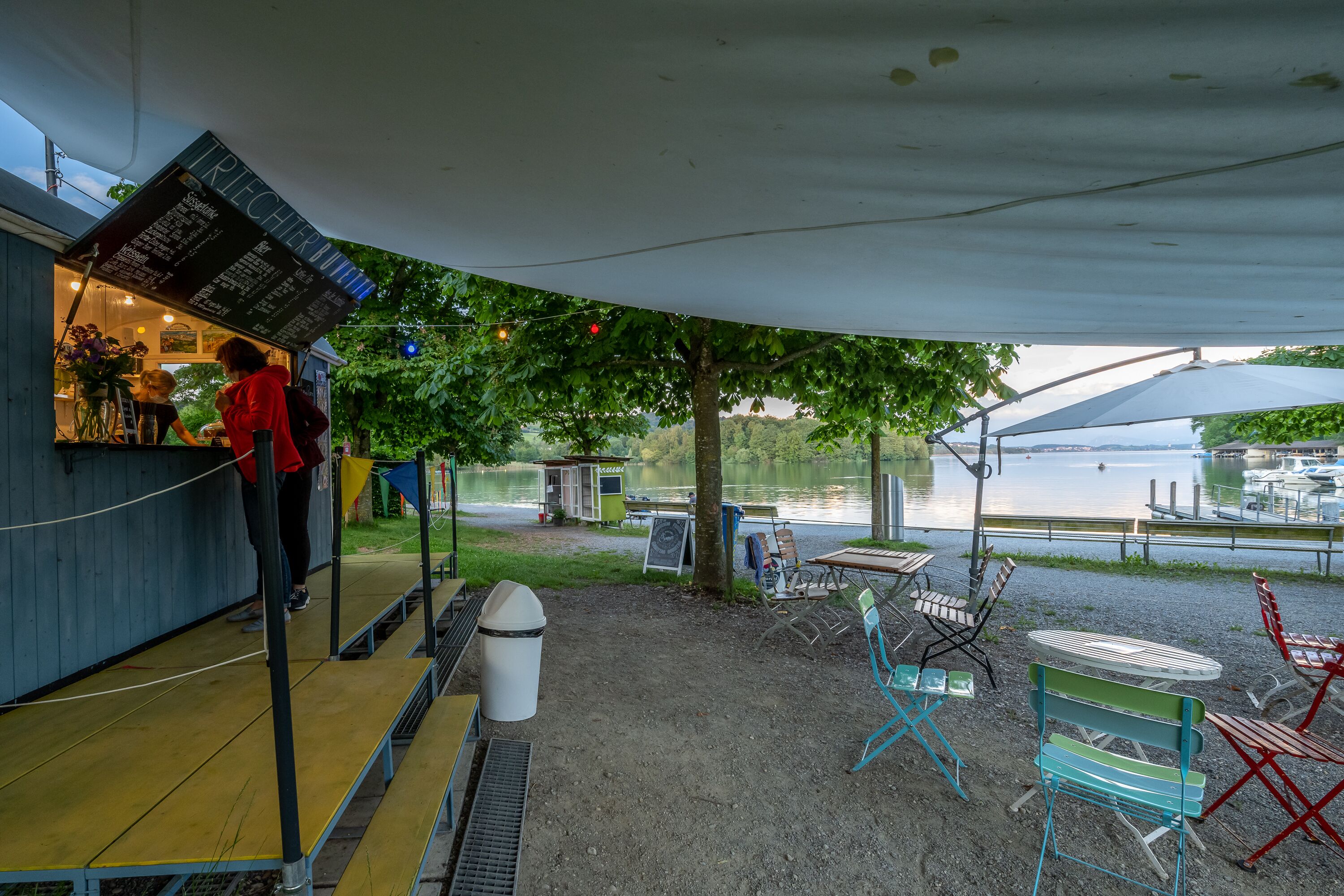 An outdoor seating area with a view of a lake, tables, chairs, and a food service counter.