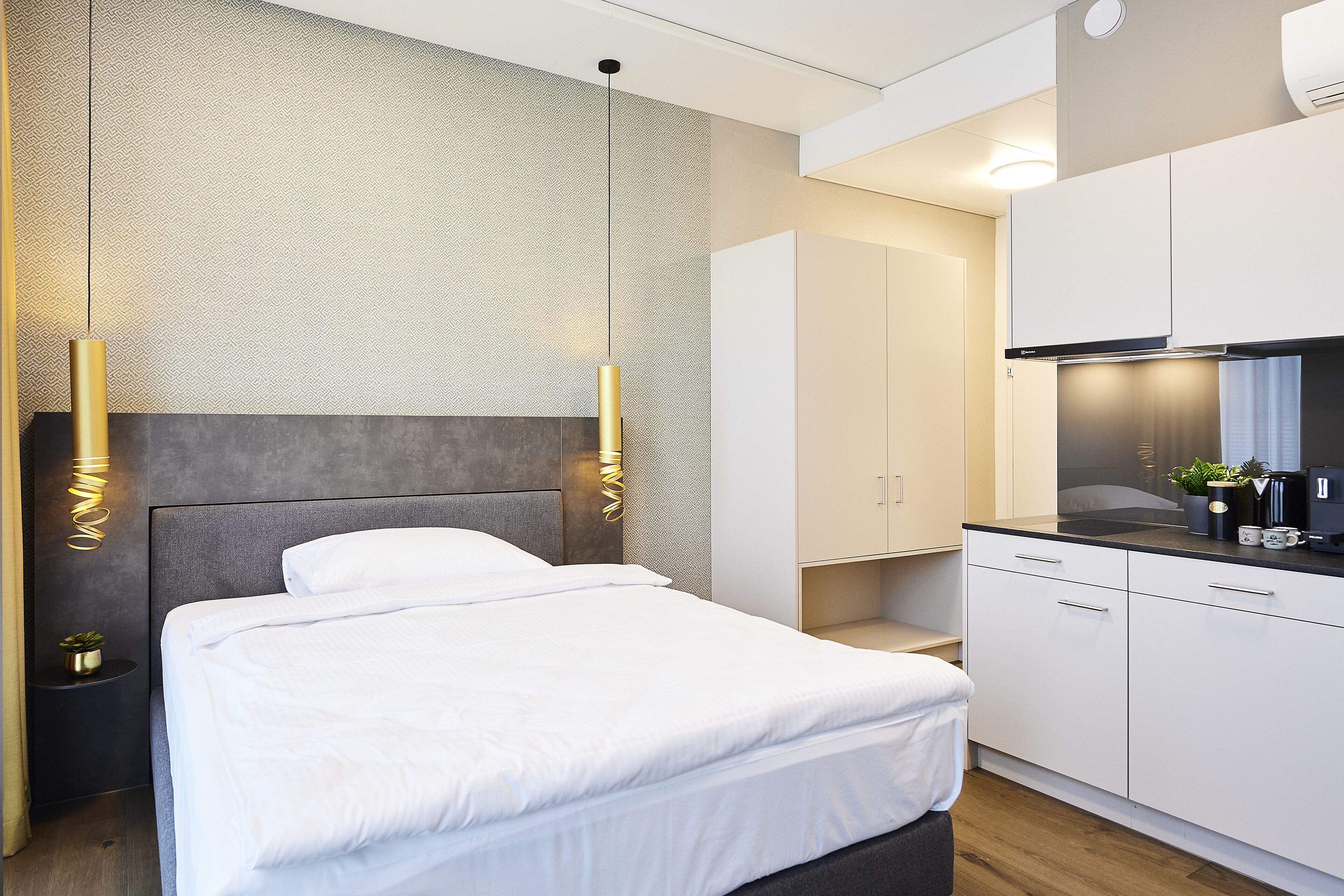 Une chambre d’hôtel moderne avec un lit bien fait, une petite table de chevet agrémentée d’une plante et une kitchenette compacte équipée d’une cuisinière et de placards.