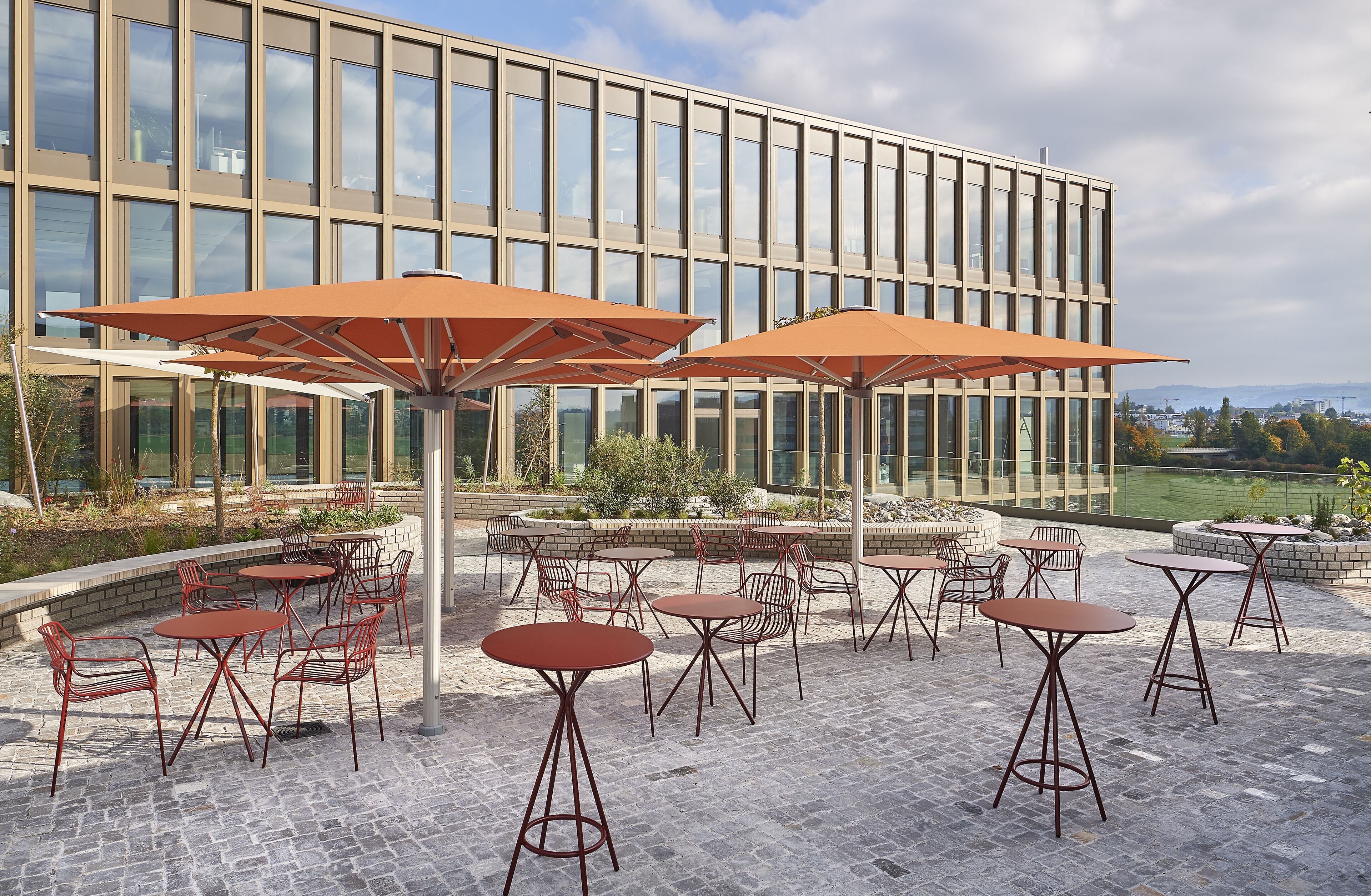 Un patio exterior con mesas y sillas, un edificio moderno al fondo y vistas a un parque y un puente.