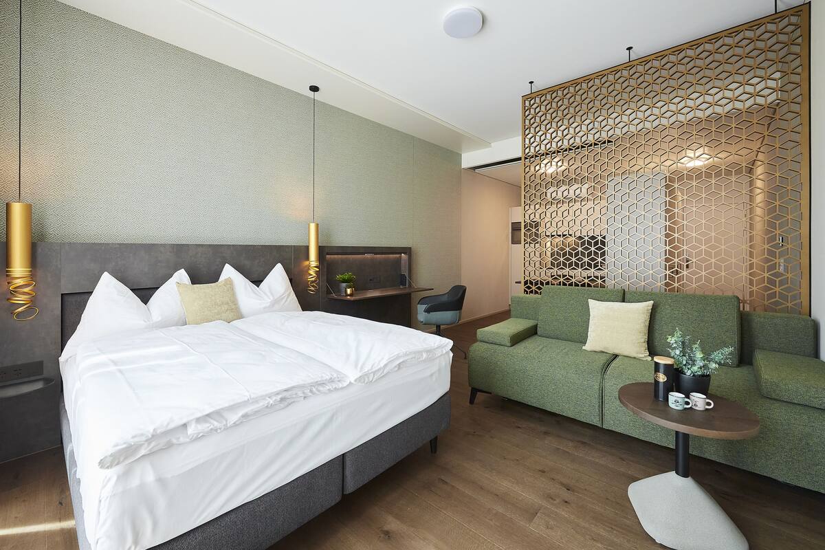 A modern hotel room with a large bed, a green sofa, a small table, and a decorative partition.