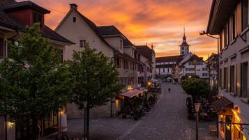 A cobblestone street lined with traditional European buildings, a church tower, and outdoor seating areas with tables and chairs.