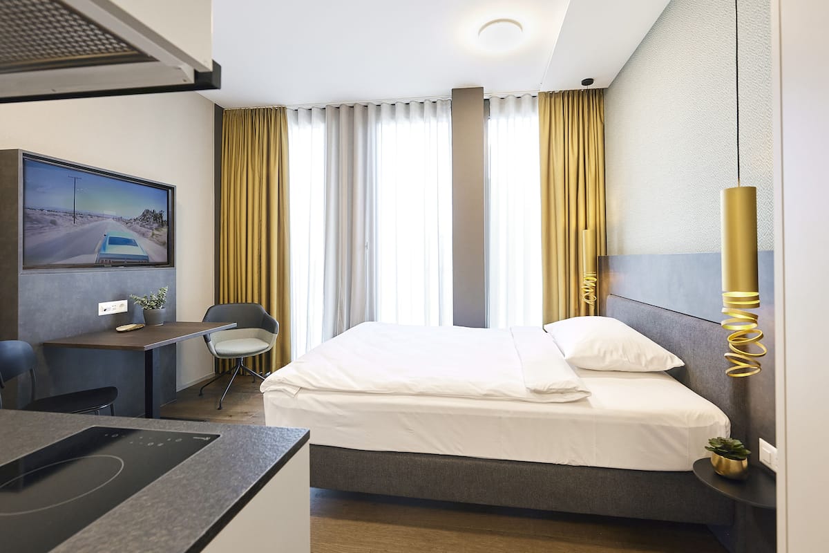 A hotel room with a bed, a desk, a chair, a TV, and a plant.