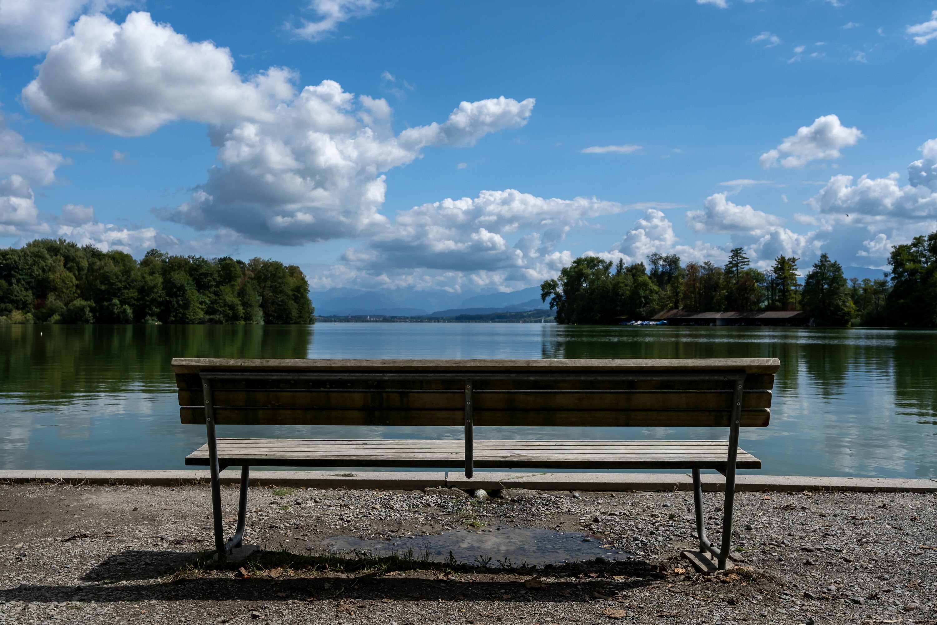 Eine Bank am See mit Bäumen und Bergen im Hintergrund.