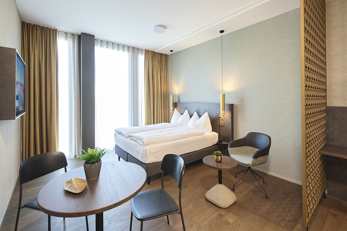 A hotel room with a bed, two chairs, a small table, a TV, and a plant.